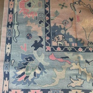 May include: A detailed close-up of a vintage rug with a complex floral pattern. The rug features a light blue central field with pink and navy blue accents. The border is a darker navy blue with geometric designs, and the overall aesthetic is a blend of traditional and ornate.