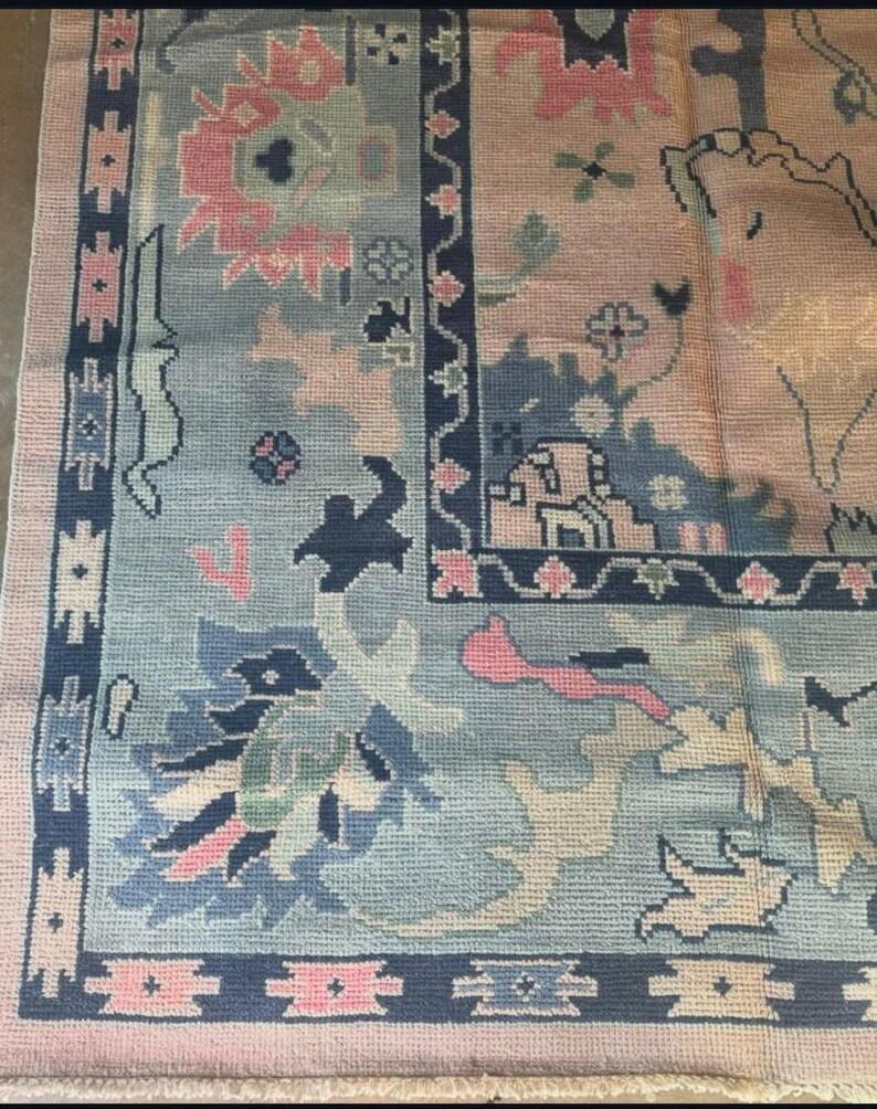 May include: A detailed close-up of a vintage rug with a complex floral pattern. The rug features a light blue central field with pink and navy blue accents. The border is a darker navy blue with geometric designs, and the overall aesthetic is a blend of traditional and ornate.
