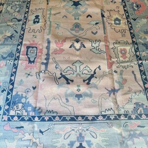 May include: A large, rectangular rug with an intricate, traditional design. The rug features a central field in a muted peach color, adorned with floral and geometric motifs in shades of blue, pink, and green. A dark blue border frames the design.