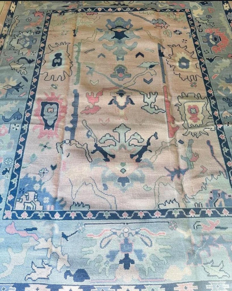 May include: A large, rectangular rug with an intricate, traditional design. The rug features a central field in a muted peach color, adorned with floral and geometric motifs in shades of blue, pink, and green. A dark blue border frames the design.