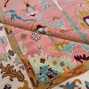 May include: A close-up of a pink rug with an intricate pattern. The rug features a combination of colors, including pink, teal, gold, and purple. The design includes floral and geometric shapes, with a rolled-up section revealing the rug's texture.