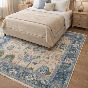 May include: A bedroom scene featuring a beige bed with white bedding and decorative pillows. A patterned rug with blue, pink, and green floral designs sits on the wooden floor. Two bedside tables with lamps flank the bed.
