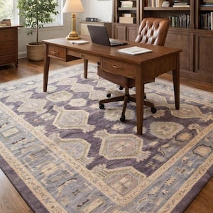 May include: A home office featuring a wooden desk with a laptop and lamp, a leather chair, and a large patterned rug in shades of purple, blue, and beige. A built-in bookcase and a potted plant are also visible.