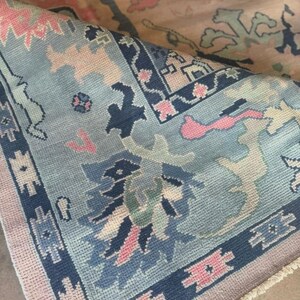 May include: A detailed shot of a folded rug with a light blue and pink color scheme. The rug features intricate floral and geometric patterns in shades of blue, pink, and cream. The edges are bordered with a dark blue design.