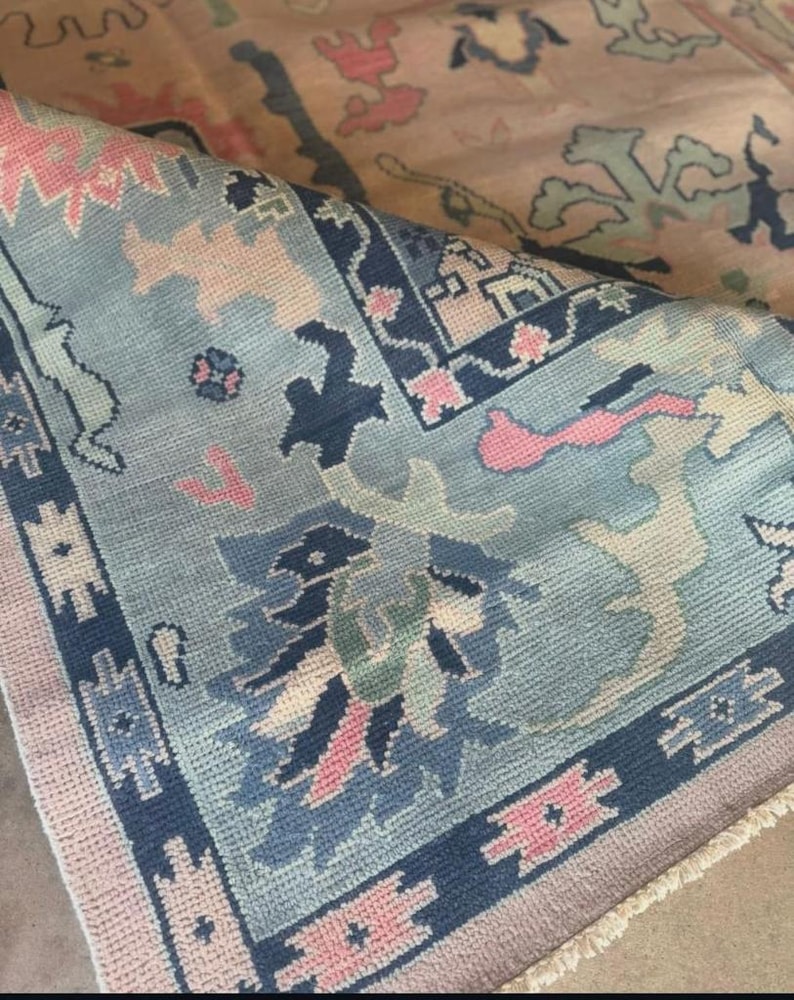 May include: A detailed shot of a folded rug with a light blue and pink color scheme. The rug features intricate floral and geometric patterns in shades of blue, pink, and cream. The edges are bordered with a dark blue design.