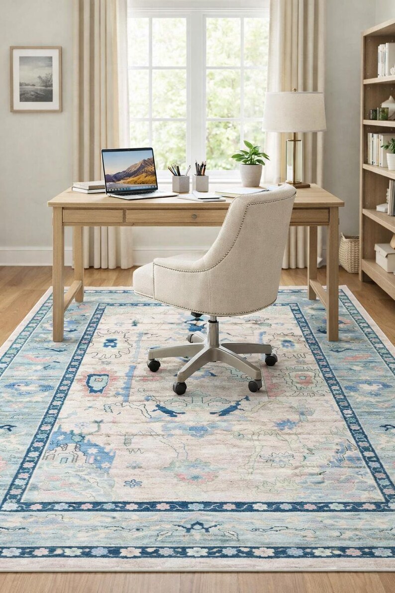 May include: A light-colored home office setup featuring a wooden desk with a laptop, pens, and a small potted plant. A beige office chair sits on a patterned rug with blue and pink accents. A bookshelf and window are in the background.