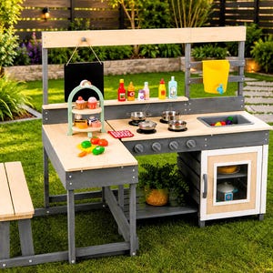 May include: A wooden outdoor play kitchen featuring a grey and natural wood finish, a sink, a stove with four burners, a side bench, a chalkboard, a yellow water dispenser, and various toy food accessories including cupcakes and plastic vegetables.