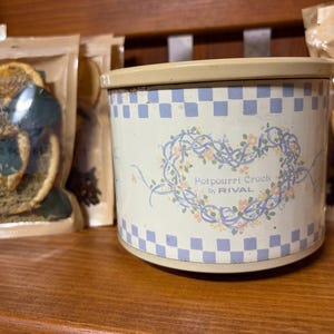 May include: A cylindrical cream-colored metal potpourri crock by RIVAL with a blue and white checkered border at the top and bottom. The center features a blue vine heart design with small orange flowers and the text Potpourri Crock by RIVAL.