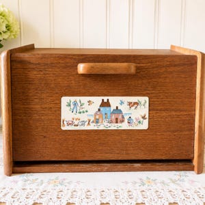May include: A rectangular wooden bread box with a drop-down front door and a horizontal handle. The center of the door features a rectangular decal depicting a rural farm scene with houses, a person gardening, a cow, pigs, and various plants.