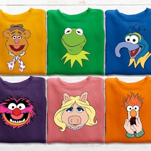 May include: Six crewneck sweatshirts in mustard yellow, green, blue, purple, dusty pink, and orange. Each features a screen-printed graphic of a Muppet character: Fozzie Bear, Kermit the Frog, Gonzo, Animal, Miss Piggy, and Beaker, respectively.