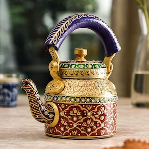 Pode incluir: Uma chaleira de metal decorativa com um padrão floral vibrante pintado à mão em vermelho, dourado, verde e azul. A chaleira possui um bico curvo, uma tampa com um pequeno botão e uma alça roxa espessa e arqueada com detalhes dourados.