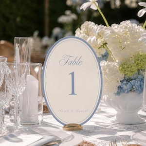 May include: An oval table number card featuring a light blue border and the text Table 1 with the names Sarah & Daniel printed in a script font. The card is held upright by a small gold-colored circular base on a white patterned tablecloth.