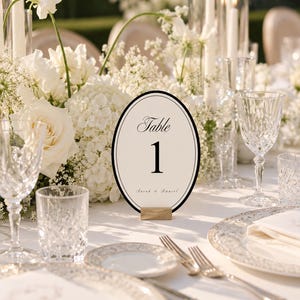 Puede incluir: Una tarjeta ovalada de número de mesa con fondo crema, borde negro, el texto "Table 1" en fuente serif y los nombres "Sarah & Daniel" en cursiva. La tarjeta se mantiene en posición vertical mediante un pequeño soporte rectangular de metal dorado.
