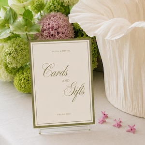 May include: A cream-colored, pleated fabric bowl with a ruffled edge sits next to a rectangular sign with a green border. The sign features the text "Olivia & Daniel", "Cards AND Gifts", and "Thank you!" surrounded by pink and green hydrangea flowers.