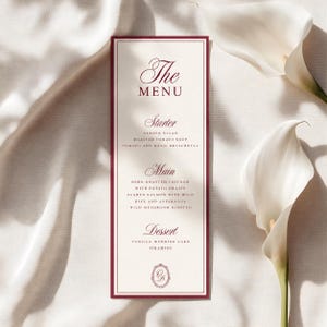 May include: A rectangular menu card with a deep red border on a cream background. The text reads The Menu, Starter, Garden Salad, Roasted Tomato Soup, Tomato and Basil Bruschetta, Main, Herb-Roasted Chicken, Seared Salmon, Wild Mushroom Risotto, Dessert, Vanilla Wedding Cake, Tiramisu.