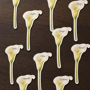 May include: Calla lily place cards with names printed on them. The white and yellow flowers are arranged on a dark brown wooden surface. Each card has a different name written in black script.