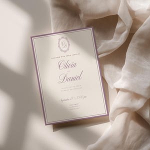 Puede incluir: Una tarjeta de invitaci&oacute;n de boda rectangular con fondo color crema y un borde fino de color morado. El texto dice Together with their families, Olivia and Daniel, invite you to their wedding celebration, September 17, 7:00 pm, con un monograma.