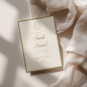 May include: A wedding invitation with a cream background and olive green border. The invitation features the names "Sarah" and "Daniel" in elegant script, along with event details. The invitation is set against a soft, draped fabric.