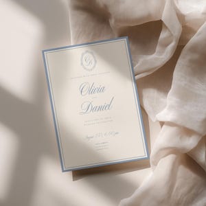 May include: A rectangular wedding invitation card with a cream-colored background and a dusty blue border. The text reads Olivia and Daniel, invite you to their wedding celebration, August 25, 6:00 pm, with a decorative monogram at the top.