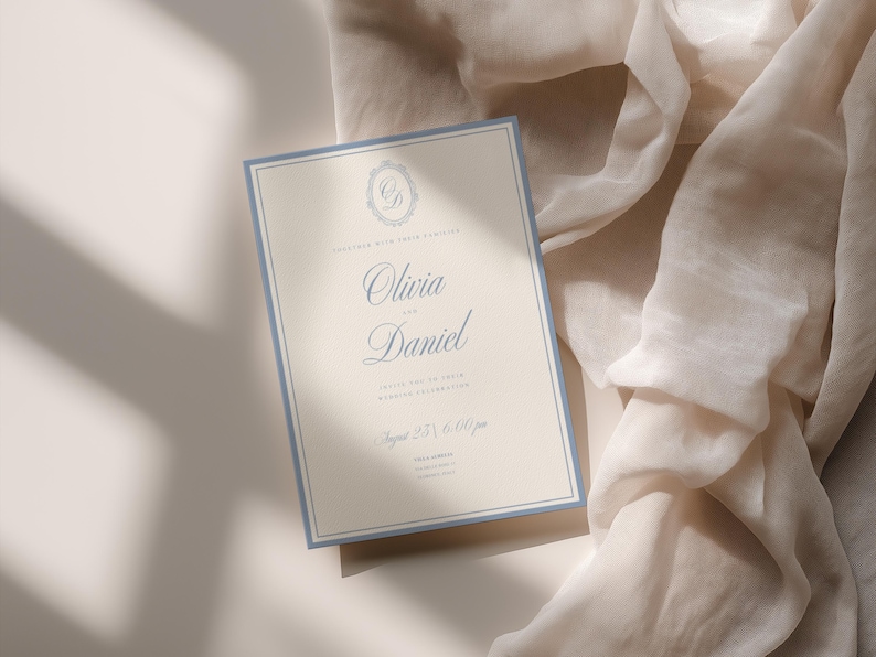 May include: A rectangular wedding invitation card with a cream-colored background and a dusty blue border. The text reads Olivia and Daniel, invite you to their wedding celebration, August 25, 6:00 pm, with a decorative monogram at the top.
