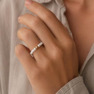 Può includere: Un anello caratterizzato da una serie di piccole perle bianche rotonde disposte a fascia, con una singola sfera color oro lucido posizionata al centro. Le perle sono uniformi per dimensioni e colore, creando un design circolare continuo.