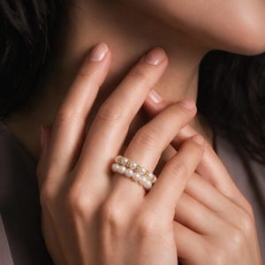 Op de afbeelding: Een dubbele ring met twee rijen kleine, ronde, witte parels gescheiden door een dunne, getextureerde goudkleurige metalen band. De ring wordt om een vinger gedragen en toont het afwisselende patroon van de witte parels en de goudkleurige accenten.