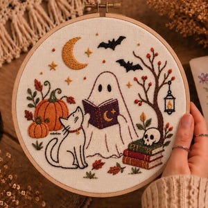 Pode incluir: Um bastidor de bordado circular com fundo de tecido branco apresentando uma cena costurada à mão com um fantasma lendo um livro roxo, um gato branco, abóboras laranja, uma caveira sobre livros, uma lanterna, morcegos e uma lua crescente.