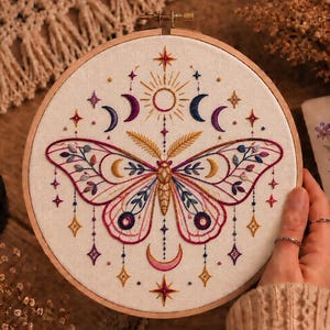 May include: A circular wooden embroidery hoop containing a cream-colored fabric panel. The fabric features a detailed hand-stitched moth design in shades of pink, blue, and gold, surrounded by celestial motifs including stars, suns, and crescent moons.
