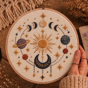 Puede incluir: Un bastidor de bordado circular con un diseño celestial cosido a mano sobre tela color hueso. El centro muestra un sol dorado rodeado de varios planetas, una gran luna creciente azul oscuro, fases lunares y estrellas doradas.