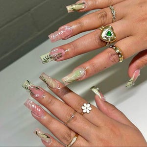 Puede incluir: Primer plano de uñas manicuradas con varios diseños. Algunas uñas son de color rosa claro con un acabado transparente y brillante, mientras que otras presentan acentos verdes y beige con líneas doradas y detalles de perlas. Varios anillos de oro con piedras preciosas y diseños florales adornan los dedos.