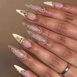 May include: Close-up of a hand with long, almond-shaped nails. The nails are painted with a light pink base, white tips, and gold embellishments. Some nails feature gold frames and small jewels.