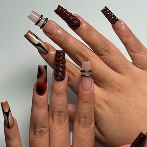 Op de afbeelding: Een close-up van een hand met lange, gebeeldhouwde acrylnagels. De nagels hebben verschillende ontwerpen, waaronder een wervelpatroon, een stippenpatroon en een gradiëntontwerp. Sommige nagels hebben zwarte ringaccenten. Het kleurenpalet omvat tinten bruin, beige en zwart.