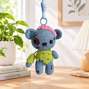 May include: A blue crocheted bear ornament with a pink hat, a lime green tattered shirt, and a brown button eye. The bear features black stitched details and is attached to a blue cord with a plastic ring for hanging purposes.
