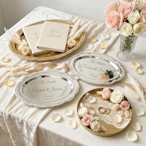 Könnte beinhalten: Zwei cremefarbene Gelübdebücher auf einem goldenen ovalen Tablett, zwei silberne ovale Tabletts mit der Gravur The Smiths, Emma & James, N.Y. New York, 12. Oktober 2026, und ein goldenes rundes Tablett mit Spitze, zwei Ringen sowie rosa und weißen Rosen.