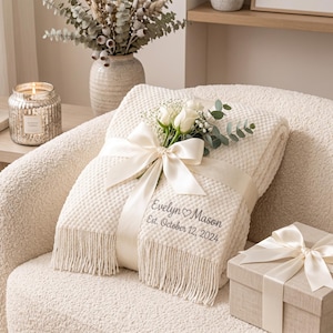 Puede incluir: Una manta de color crema con textura y flecos, atada con una cinta de satén ancha y decorada con rosas blancas. La manta está bordada con el texto "Evelyn ♡ Mason" y "Est. October 12, 2024" en una fuente cursiva de color gris oscuro.