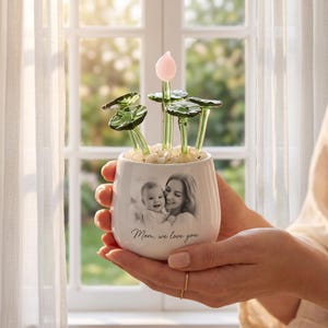 Könnte beinhalten: Ein weißer Keramikübertopf mit einem schwarz-weißen Foto eines Elternteils und eines Kindes sowie dem darunter gedruckten Text "Mom, we love you". Der Übertopf ist mit kleinen weißen Steinen und mehreren grünen Glaspflanzenstielen gefüllt.