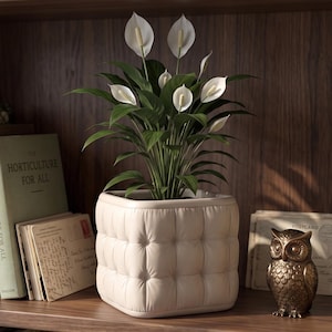 May include: A peace lily plant with white spathes and dark green leaves sits in a square, tufted, off-white ceramic planter. The planter features a grid-like, button-tufted texture on all four sides, creating a soft, cushioned appearance.