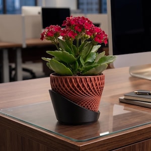 May include: A vibrant Kalanchoe plant with clusters of small red flowers and thick green leaves, potted in a textured, terracotta-coloured planter that sits inside a sleek, black, angled base with a modern, geometric, lattice-like pattern.