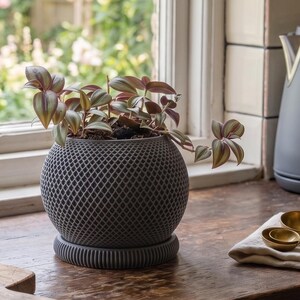 May include: A spherical dark grey planter with a textured diamond-patterned exterior and a matching circular ribbed base. The planter contains a Tradescantia zebrina plant with variegated green and purple leaves that spill over the sides.