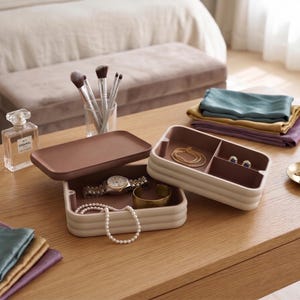 May include: A two-tiered rectangular jewellery organiser with a cream-coloured exterior and a dark brown interior. The top tray is removed and placed beside the base, which contains a wristwatch, a gold cuff, and a string of white pearls.