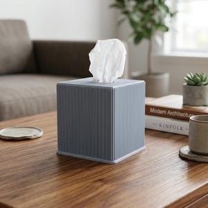 May include: A square, light grey tissue box cover featuring a vertical ribbed texture on all four sides. The cover has an open top through which a single white tissue is pulled upwards. The object has a clean, geometric shape and a matte finish.
