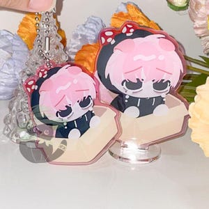 May include: Two acrylic charms featuring a cartoon character with pink hair and a black outfit. The character is wearing a black hood with a red bow. One charm is attached to a keychain, and the other is on a clear stand.