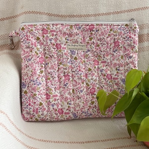 May include: A rectangular quilted zipper pouch featuring a dense floral pattern in shades of pink, purple, and green on a white background. A small rectangular fabric label is centered on the front with the text The Thrifting Therapist handmade goods.