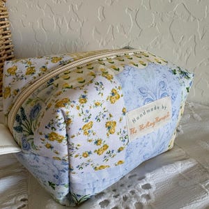 May include: A handmade, rectangular cosmetic bag with a floral patchwork design. The bag features a cream-colored zipper and a small tag that reads "Handmade by The Thrifting Therapist". The fabric includes patterns of yellow flowers and blue floral motifs.