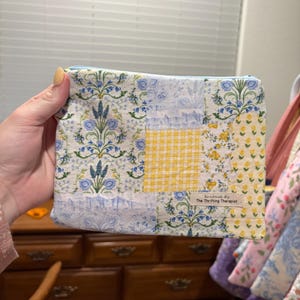 May include: A handmade patchwork zippered pouch featuring floral and gingham patterns in blue, yellow, and white. The pouch has a blue zipper and a label that reads "Handmade By The Thrifting Therapist."
