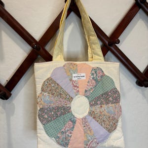May include: A quilted tote bag featuring a large, multi-colored floral and solid pastel fabric flower appliqué on a cream background. The bag has two pale yellow shoulder straps and a small white label that reads The Thrifting Therapist.