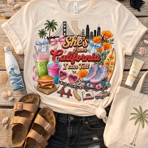 May include: A cream-colored t-shirt featuring a graphic print with the text "She's from California I can Tell," a skyline, palm trees, a burger, drinks, heart-shaped sunglasses, roller skates, a surfboard, and various California-themed icons.