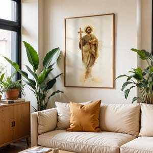 Op de afbeelding: Een ingelijste wandkunstprint met een aquarelillustratie van een bebaard figuur in vloeiende beige gewaden, die een houten kruis in de ene hand en een brood in de andere hand houdt, met een gouden aureool boven het hoofd.