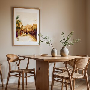 May include: A framed wall art print depicting a crowd scene with a figure on a donkey, displayed above a light wood dining table with two round ceramic vases containing green branches, and two wooden chairs with woven seats.