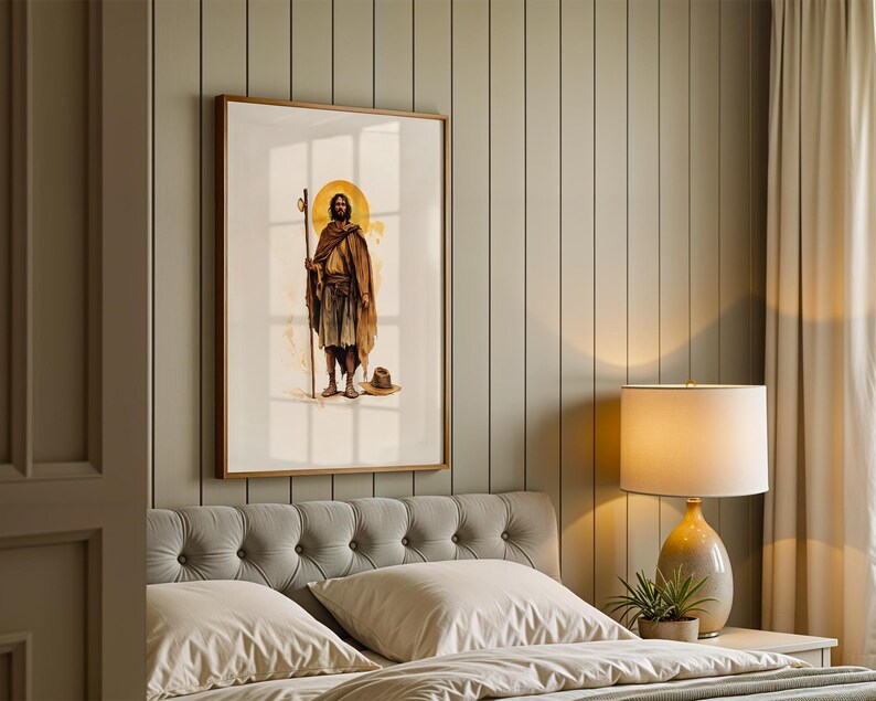 Op de afbeelding: Een ingelijste wandkunstprint met een staande figuur in bruine gewaden die een houten staf vasthoudt met een gouden halo achter het hoofd. De figuur is afgebeeld in een aquarel-stijl met een kleine hoed op de grond bij de voeten.